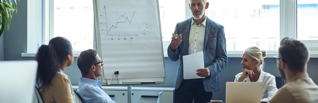 A business owner leads a meeting in a conference room, speaking to colleagues seated around a table with a flip chart showing a line graph.